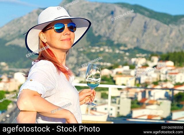 beautiful woman in straw hat enjoying evening with wine glass at resort. Luxury hotel terrace. Europe summer vacation. Happy woman drinking white wine relaxing...