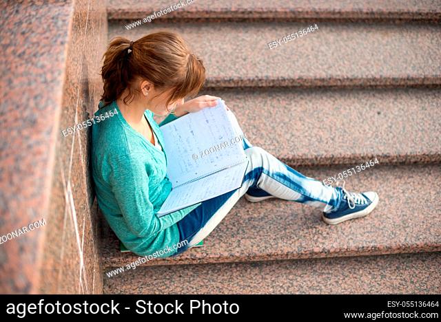 Portrait of a beautiful teenage student girl sitting on stairs in park with copy book and studing. Sunny summer day. Modern and casual lifestyle