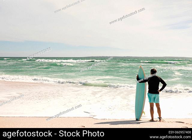 Full length of biracial senior man holding surfboard standing on shore at beach during sunny day