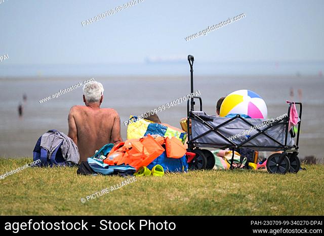 09 July 2023, Lower Saxony, Schillig: A man is sitting with two boys on a meadow in front of the bathing beach of Schllig