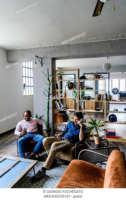 Two young men with cell phones and headphones sitting in armchairs in a loft
