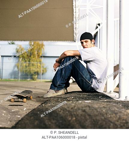 Young man with skateboard sitting outdoors, side view, portrait
