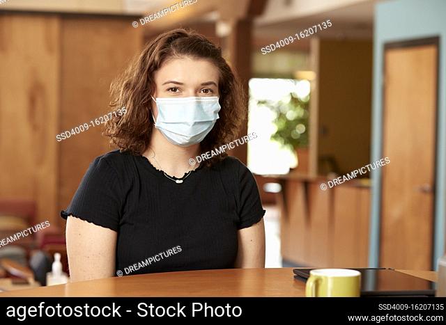 Portrait of young caucasian female wearing black t shirt and face mask, sitting at bar in kitchen of downtown loft with iPad and coffee mug