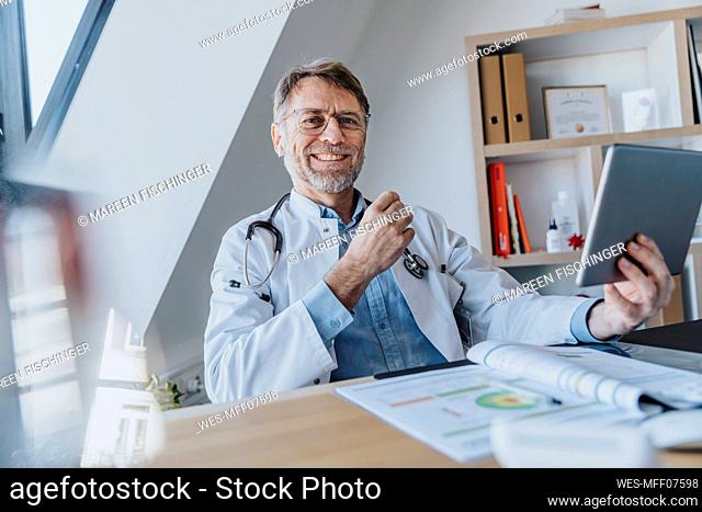 Smiling male doctor with digital tablet sitting by desk at doctor's office