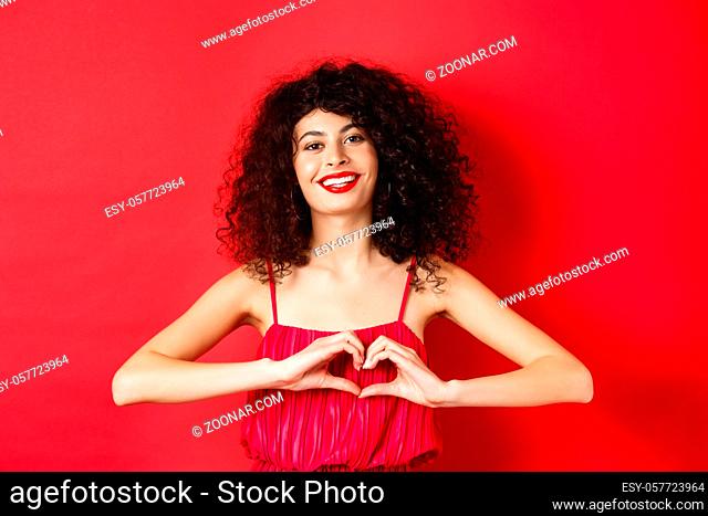 Lovers day. Beautiful woman celebrating valentines, showing heart sign and smiling, standing in romantic red dress on studio background