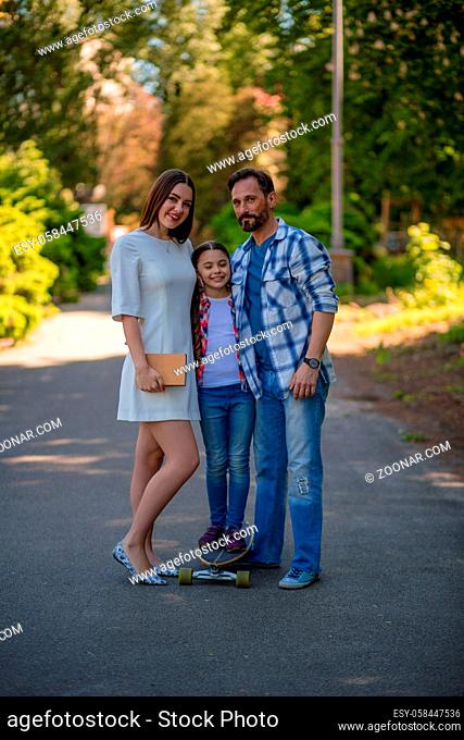 Happy Family With Kid In Park. Girl Standing On Skateboard. Green Trees On Background. Happy Family Concept