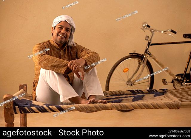 A RURAL MAN COMFORTABLY SITTING ON COT AND LOOKING AT CAMERA