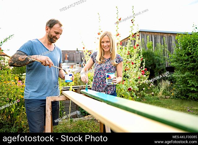 Cheerful couple painting wooden plank in garden