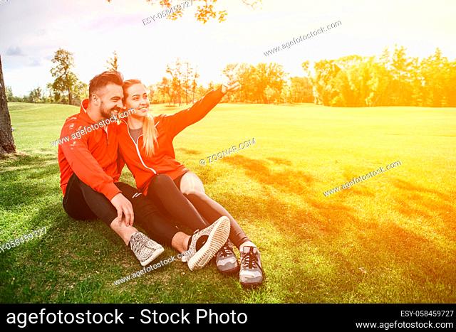 Sport man and woman making selfies in green park or forest while sitting on green grass. Happy couple hugging and posing for camera. Toned
