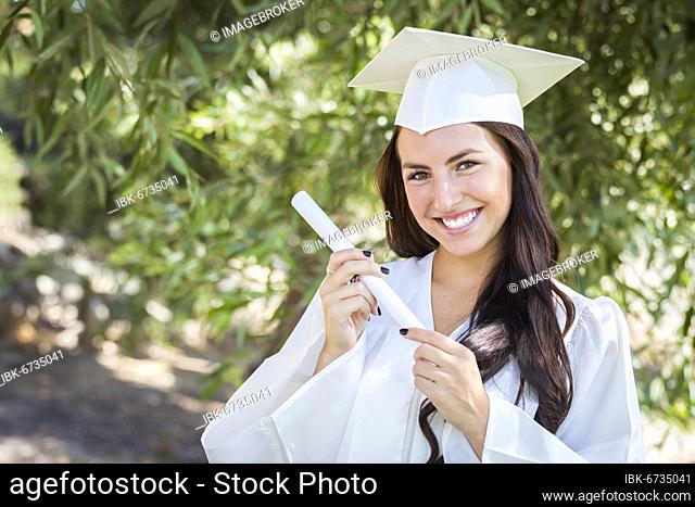 Attractive mixed-race girl celebrating graduation outside in cap and gown with diploma in hand