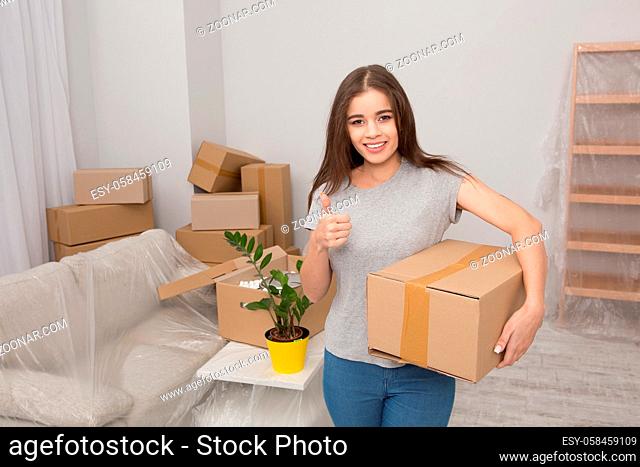 Beautiful girl moving in the new apartment. Woman after relocating standing in the room holding cardboard box and showing her thumb up