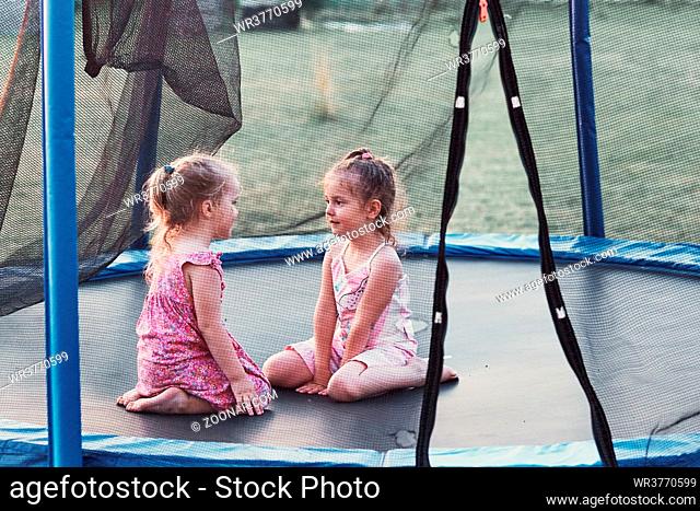 Two little cute girl talking, sitting and playing on trampoline in backyard on summer day. Candid people, real moments, authentic situations