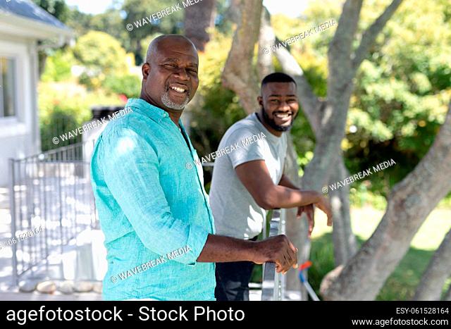 Portrait of smiling senior african american father and adult son standing on terrace in sunny garden