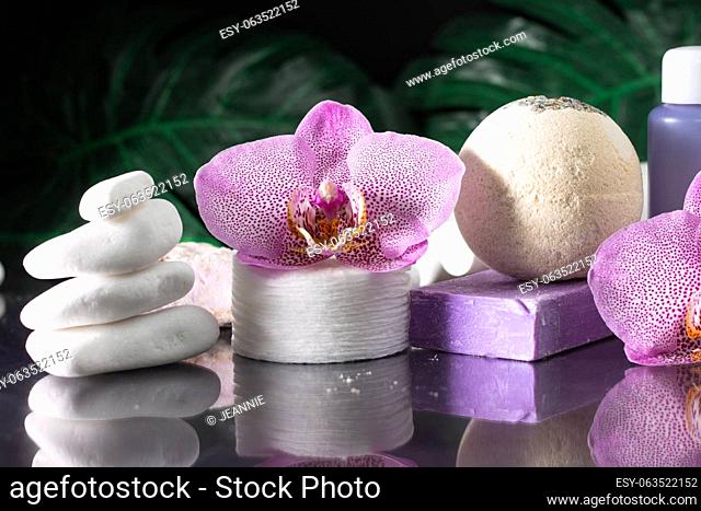 Beautiful lilac orchid flower, cosmetic bottle, cotton pads and handmade soap with stack of white stones and monstera leaves on black surface