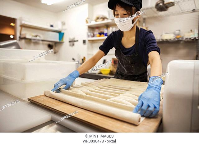 Woman working in a bakery, wearing protective gloves and mask, placing dough on large wooden board
