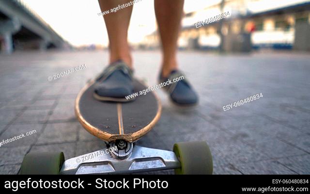 The Legs Of A Light-Skinned Man Are Gaining Speed On A Longboard On A City Street. Close-Up. Blurred Background. Bottom View