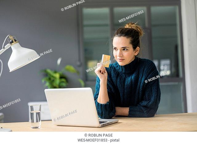 Portrait of confident woman with laptop and credit card sitting at desk at home looking at distance