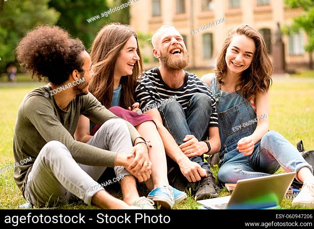 Happily laughing students on grass in University park. Group of young people sitting on lawn, having fun