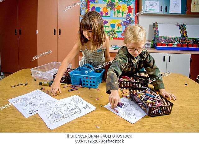 Kindergarten children in San Clemente, CA, collect and organize a table full of classroom objects including crayons and worksheets
