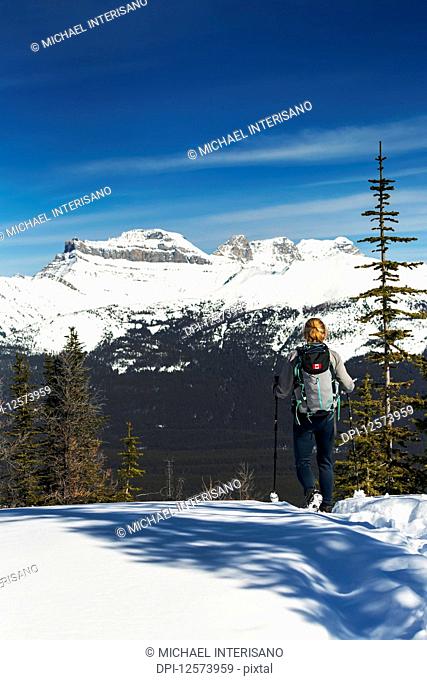 Female hiker on snow-covered pathway with snow-covered mountains, blue sky and clouds in the background; Lake Louise, Alberta, Canada