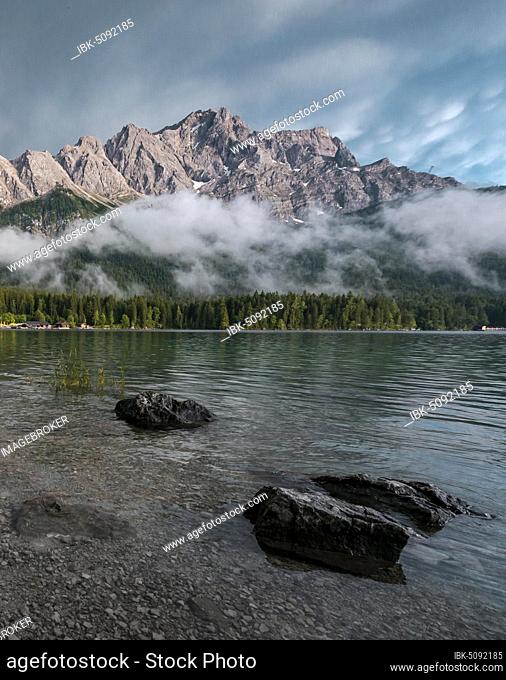 Rocks on the shore, Eibsee lake in front of Zugspitze massif with Zugspitze, low hanging clouds, Wetterstein range, near Grainau, Upper Bavaria, Bavaria