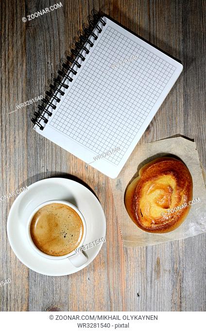 cup of espresso, checkered notebook and bun on wooden background
