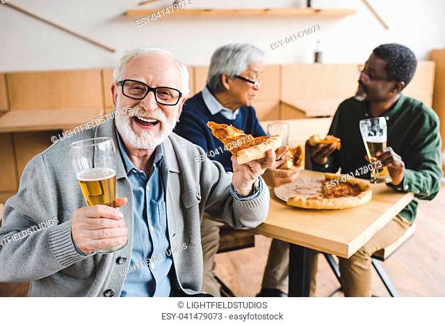 laughing senior man eating pizza with beer while his friends talking on background