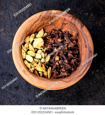 Cardamom seeds and cloves in olive wood bowl over black background. Top view. Square image