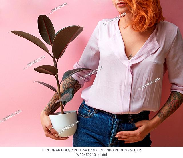 Young girl with a tattoo is holding a ficus in a flowerpot on a pink background. Mothers Day, Flowers shop concept