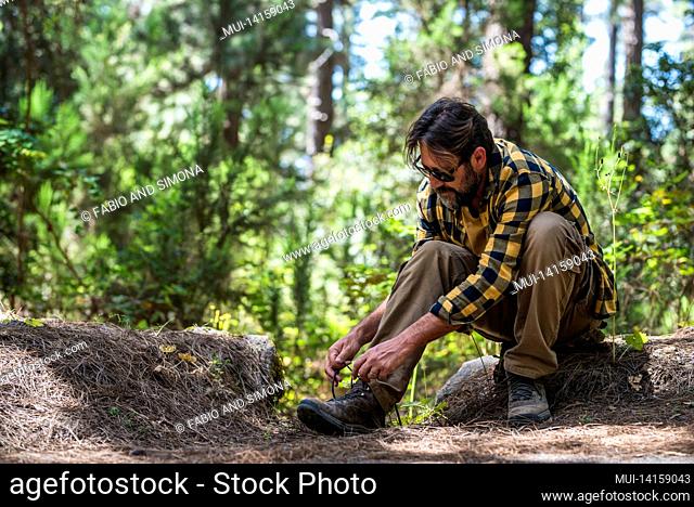 mature male hiker in sunglasses tying shoelaces and getting ready for trekking in forest while sitting on rock. man tying hiking shoelaces in forest before a...