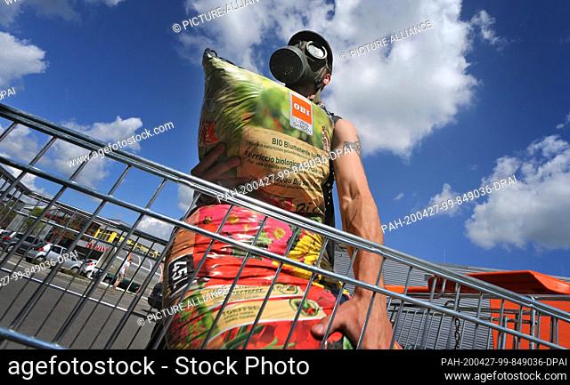 27 April 2020, Bavaria, Kaufbeuren: A young man wearing a gas mask loads the potting soil he has bought into his car in front of a hardware store