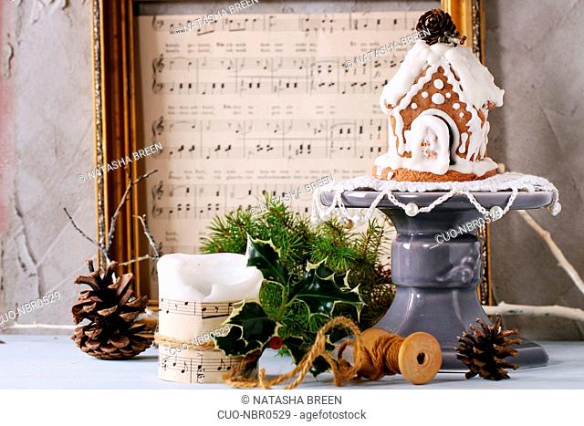 Small hand made Gingerbread house, candle and festive Christmas decor with vintage music notes over white table with plastered wall as background