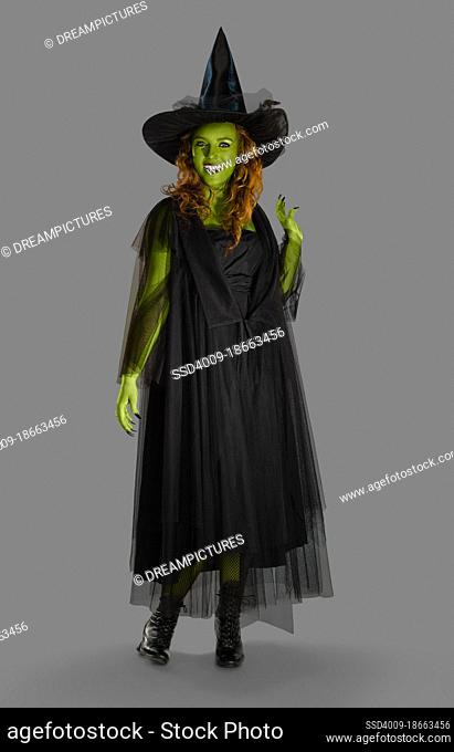 Full length portrait on gray of a woman with green body paint dressed up in a witch costume for Halloween smiling at camera