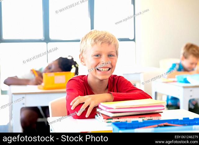 Portrait of smiling caucasian elementary schoolboy with books sitting at desk in classroom