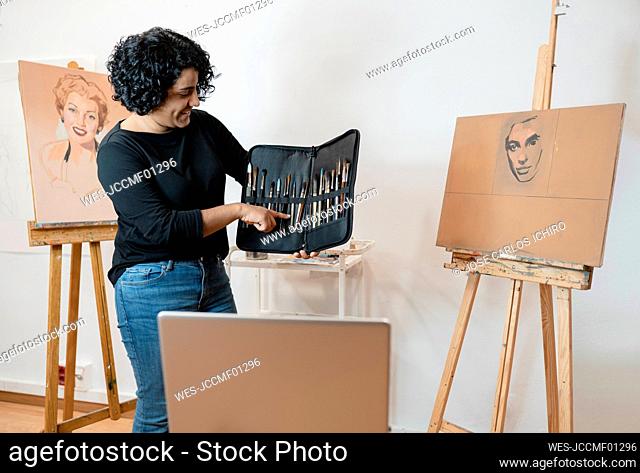 Smiling female painter standing amidst easels while pointing at paintbrush in art studio