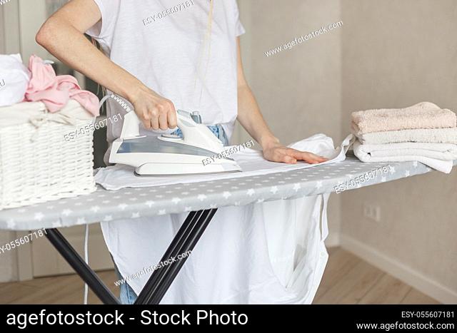 Woman ironing white clead washed shirt at home. Wicker basket and stack of color towels on ironing board