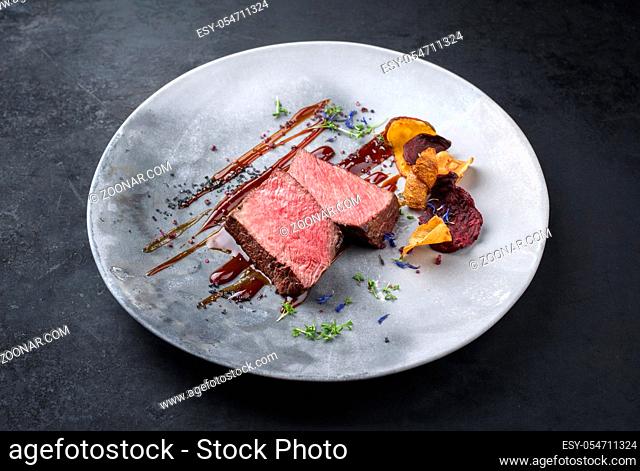 Barbecue dry aged wagyu roast beef natural sliced and offered with vegetable chips and herbs as closeup on a modern design plate with copy space