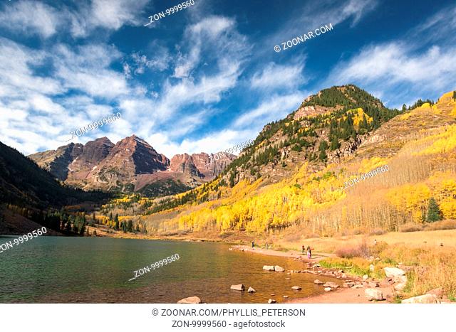 Maroon Bells Mountains are shrouded in the yellow Aspens of Fall. A small lake is at the base of the mountain peak. The sky is filled with clouds
