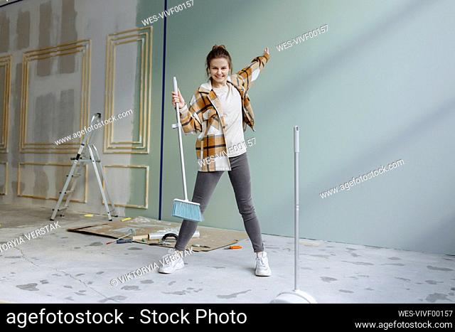 Cheerful woman in plaid shirt dancing with broom in apartment