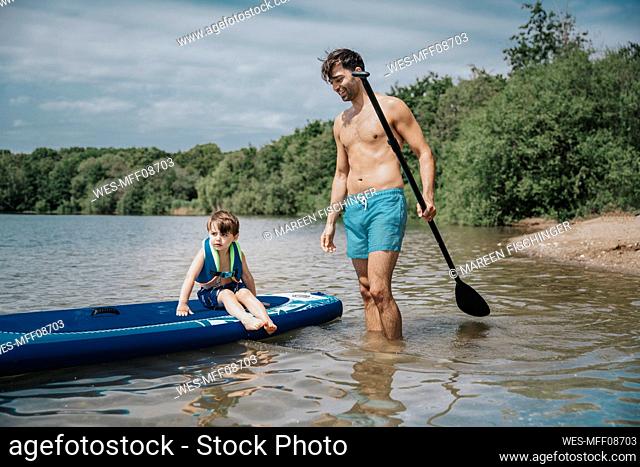 Son sitting on paddleboard and father standing in lake