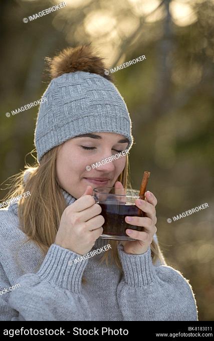 Portrait of a young woman in winter with cap, laughing, warming herself with a cup of tea, in the forest, Upper Bavaria, Bavaria, Germany, Europe