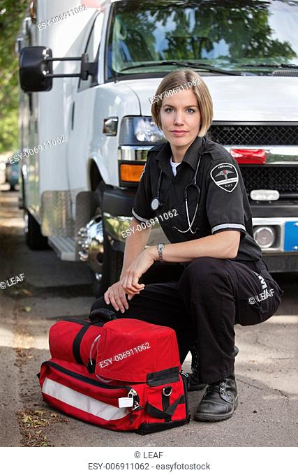Confident EMS paramedic kneeling by portable oxygen unit and ambulance