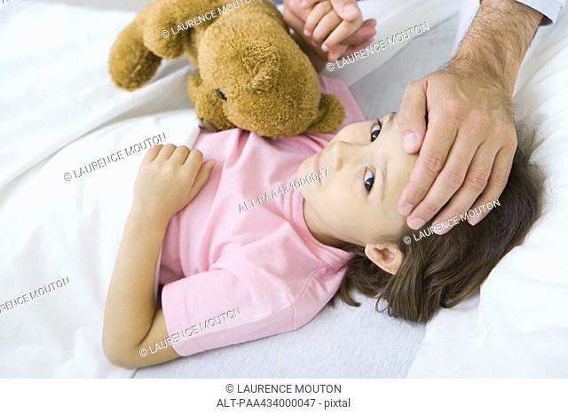 Girl lying in bed with teddy bear, man holding her hand on putting other hand on her forehead