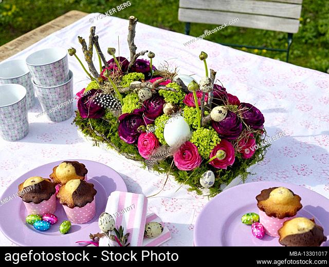 Easter breakfast table outdoors, in preparation, with coffee mugs, a stack of plates, muffins with chocolate Easter eggs on plates and a lush bouquet of flowers...