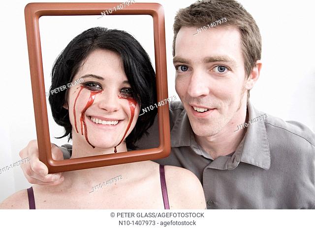 Man holding frame in front of wife, who has blood dripping from her eyes