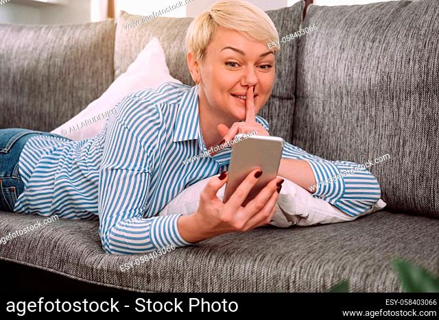 Young woman showing secret gesture while talking online by phone while lying on sofa at home