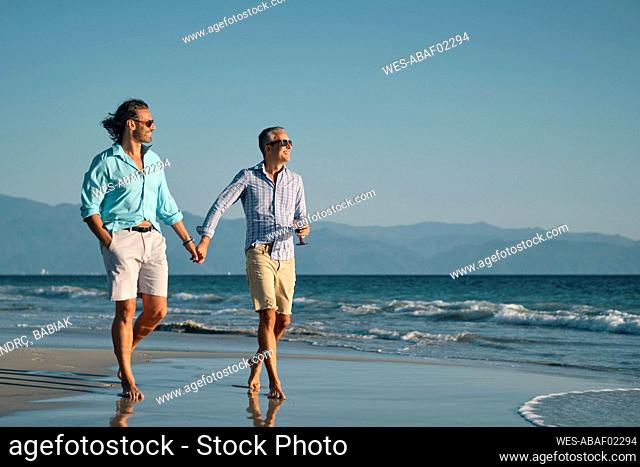 Full length of smiling mature gay men holding hands while walking on shore at beach against clear blue sky, Riviera Nayarit, Mexico
