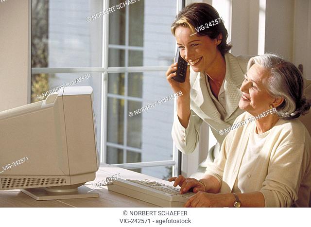 indoor, ca. 30-year-old woman stands beside her mother sitting at the table near the window at the computer making a phone call  - GERMANY, 04/03/2005
