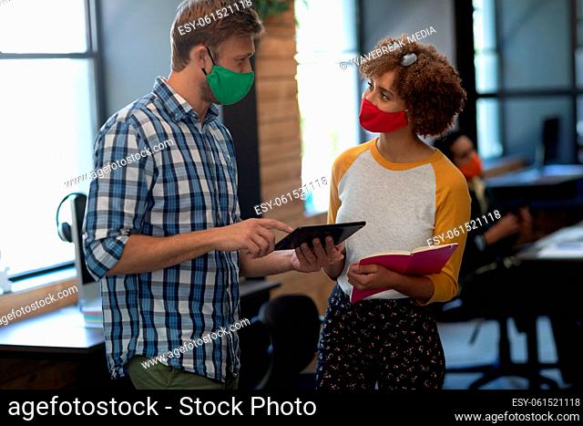 Two diverse creative colleagues wearing face masks talking in office