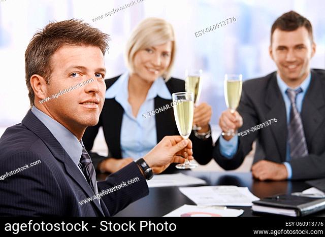 Happy middle-aged businessman sitting at meeting table at office celebrating success with champagne, smiling